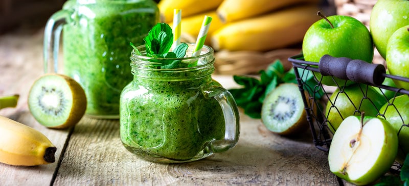 green smoothie sitting on wooden table surrounded by fruits